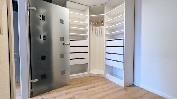 Ein begehbarer Kleiderschrank mit offenen Regalen und einer Kleiderstange, Holzboden und eine teilweise geöffnete Tür.