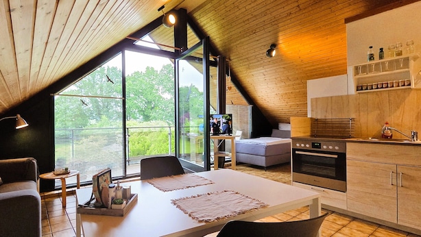 Een moderne keuken met een inbouwoven, een eethoek met een tafel en stoelen, en grote ramen met uitzicht op groen.