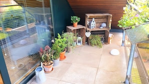 Een klein balkon met potplanten, een houten plankje met decoratieve spullen, en een glazen deur.
