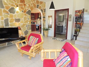 A living room with a stone wall, a television, two wicker chairs with colorful cushions, a kitchen in the background, and a staircase with a bookshelf. - Near Kalives. 2 bedrooms, private pool, Free WIFI, A.C, views and all amenities. (Chania)