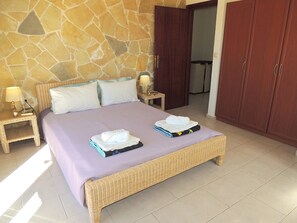 Quarto com parede de pedra, cama com colcha roxa, duas mesas de cabeceira com luminárias e um guarda-roupa.