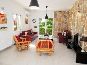 A living room with a sofa, a wooden chair, a television, and a stone wall. - Near Kalives. 2 bedrooms, private pool, Free WIFI, A.C, views and all amenities. (Chania)