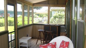 A covered porch with a wicker chair and a table, a view of the garden, and a window with a view of the outside.