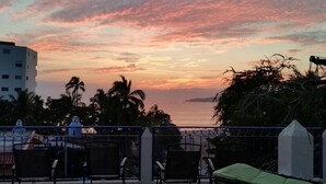 A terrace with chairs and a green lounge chair, overlooking a sunset over the sea with palm trees and a building in the background.