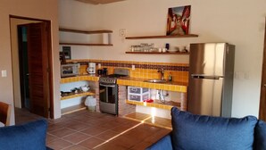 A compact kitchen with a stove, microwave, and refrigerator. There are shelves above the counter holding dishes and a painting above the sink.