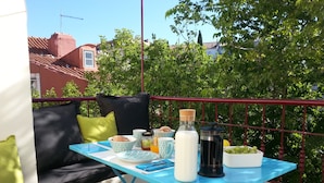 A balcony with a blue table set for breakfast, featuring a bottle of milk, a coffee pot, and a bowl of grapes.