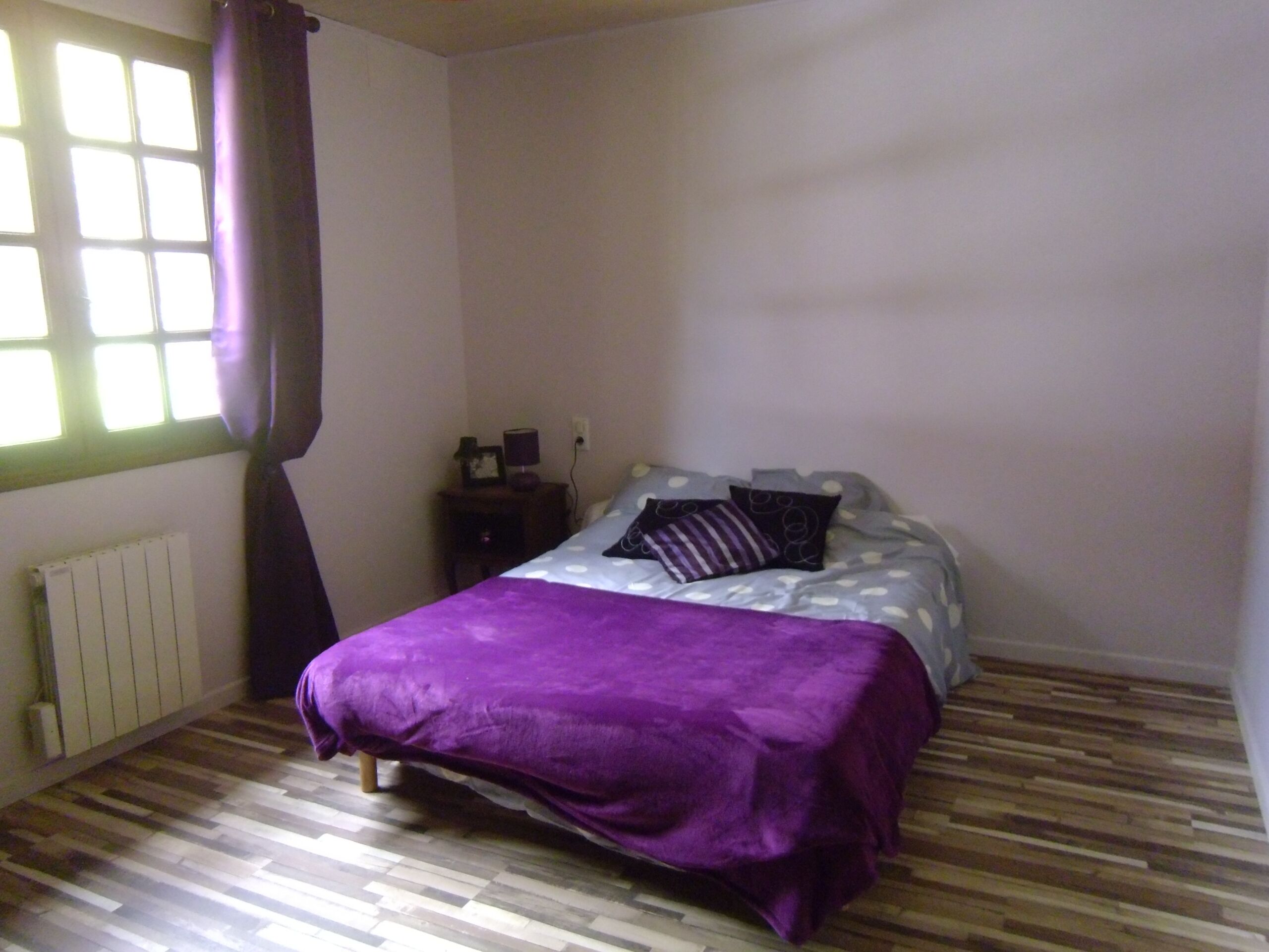 Um quarto com cama, mesa de cabeceira, janela com cortinas e radiador.