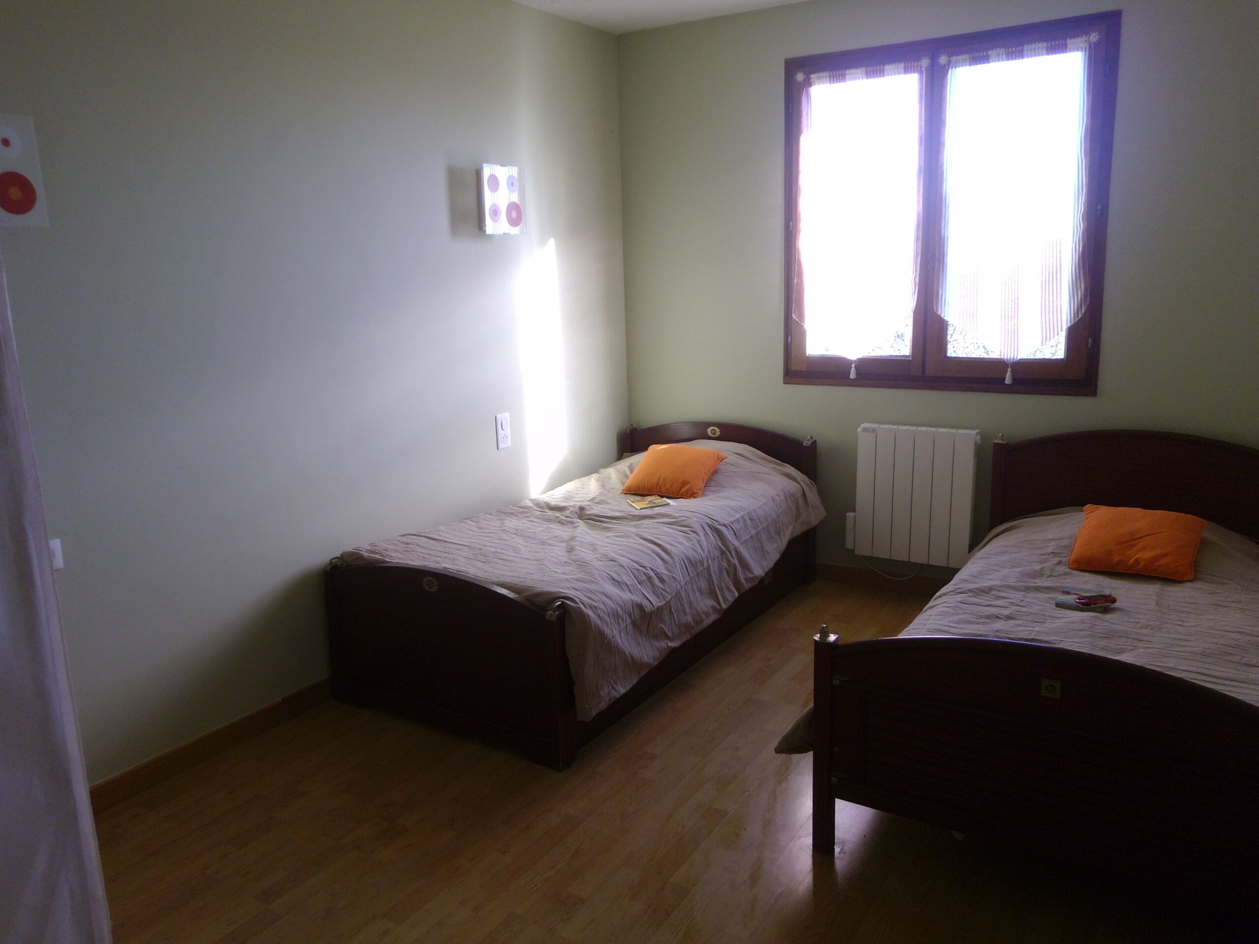 A small, well-lit room with two single beds, a radiator, and a window with curtains.