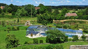 A greenhouse with a transparent roof, surrounded by a stone wall and a wooden fence, set in a lush green landscape with trees and a clear sky. - La Grange aux Lumières - Heated swimming pool - Nature (Courcy)
