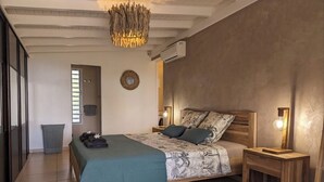 Ein Schlafzimmer mit Bett, Nachttischen, einer Hängeleuchte, einer Wandklimaanlage und einer Tür zu einem anderen Raum.