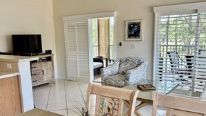 Una sala de estar con televisor, sofá, mesa de comedor con sillas y ventana con persianas.