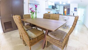 A dining area with a wooden table and wicker chairs, a kitchen in the background, and a painting on the wall. - RELAXING OCEAN VIEW CONDO (Oranjestad West)