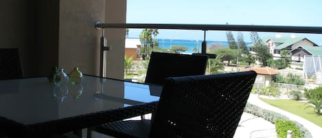 Un balcone con un tavolo in vetro e sedie in vimini, affacciato su una zona residenziale con case e cielo sereno.