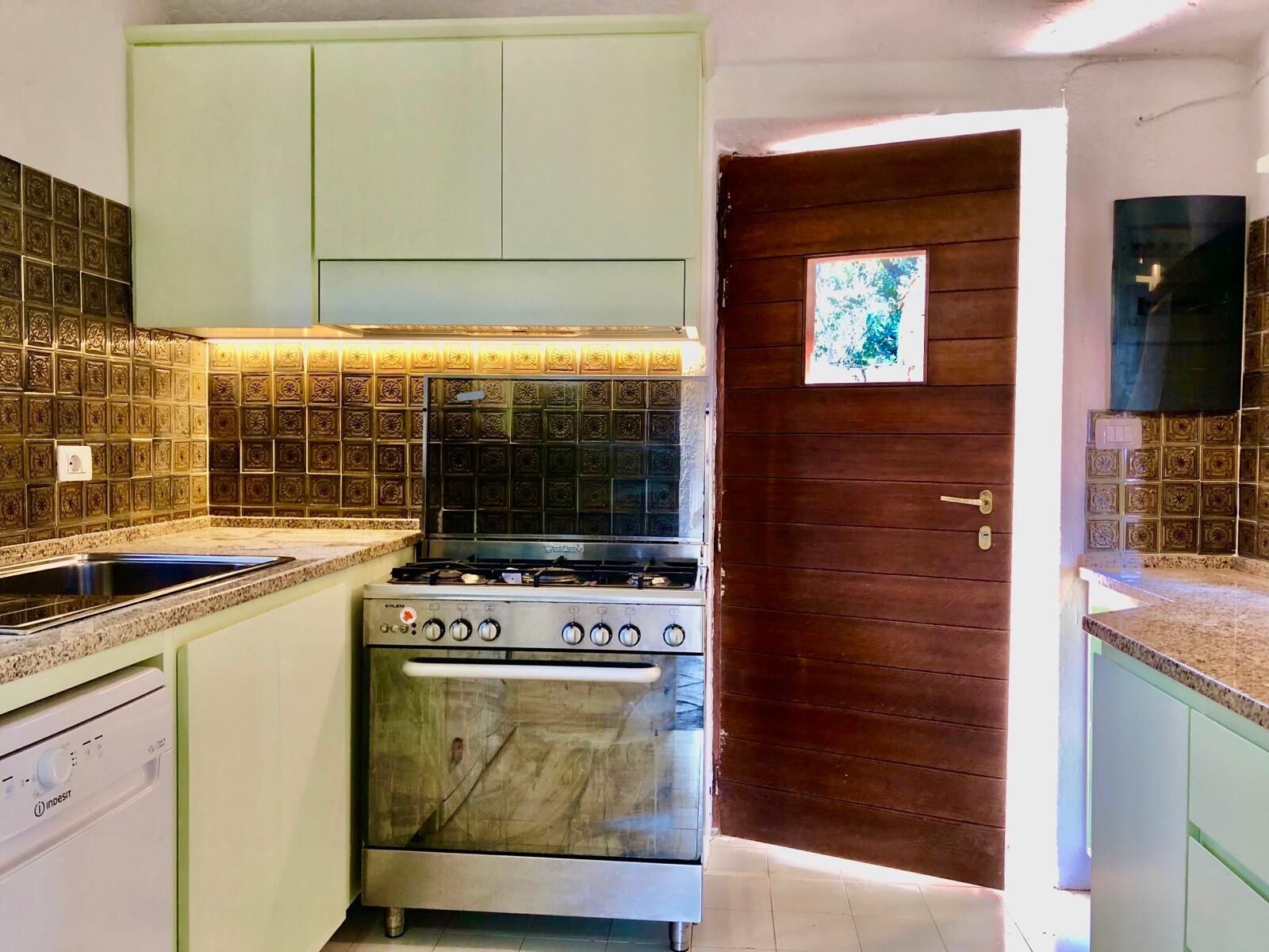 A kitchen with a stove, oven, and dishwasher. There is a wooden door with a picture on it. The walls have decorative tiles.