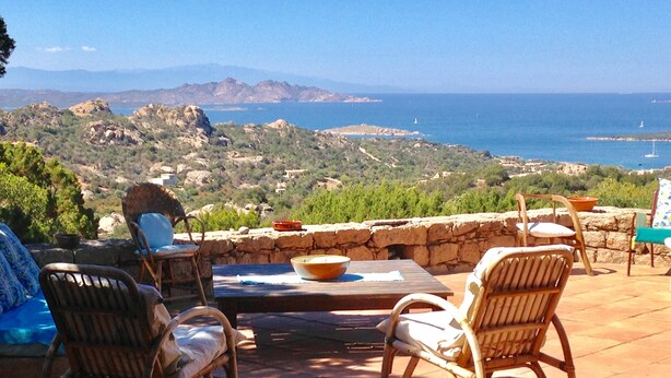Un espacio al aire libre con sillas de mimbre y una mesa, con vistas a un paisaje pintoresco con montañas y el mar.