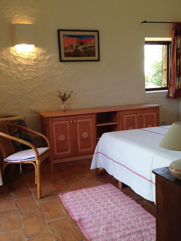 A bedroom with a bed, a wooden dresser, a chair, a small table, a window with curtains, and a painting on the wall.