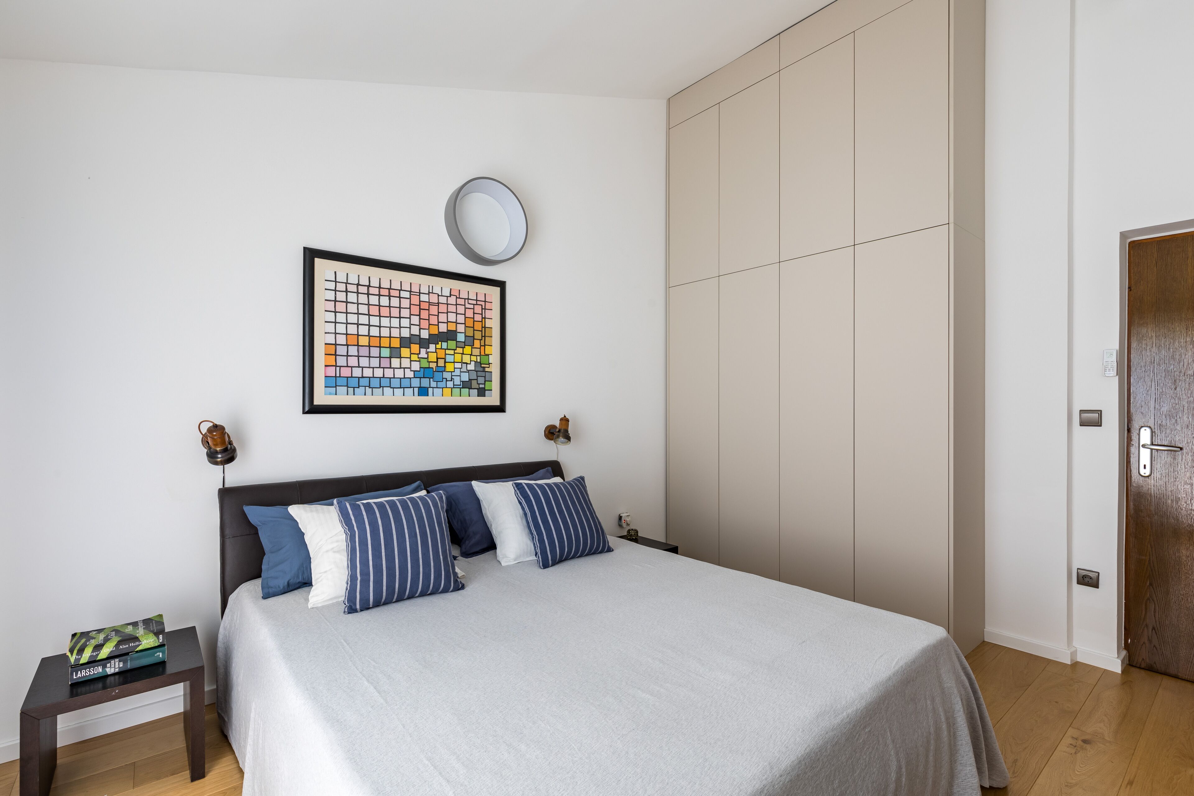 Ein ordentlich eingerichtetes Schlafzimmer mit einem Bett, einem Nachttisch mit Büchern, einem Wandbild und einem Kleiderschrank.