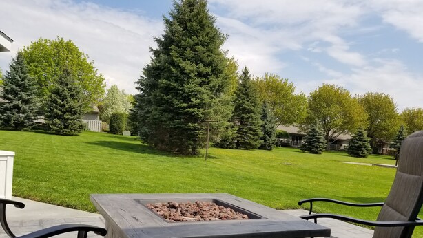 A patio with a square fire pit table and two chairs, surrounded by a well-maintained lawn and tall trees.