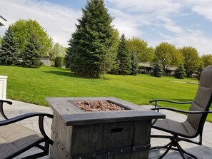 Un patio con una mesa de fogata cuadrada y dos sillas, rodeado de un césped cuidado y árboles altos.