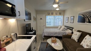 A compact living space with a kitchenette, a bed with patterned bedding, a brown sofa with star pillows, a ceiling fan, and a window with blinds. - Small Private Apartment Within Walking Distance to Beach, Restaurants & Shops (Corona del Mar)