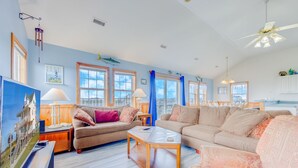 A spacious living room with a large sofa, a coffee table, and a television. There are two windows with blue curtains and a ceiling fan. - Oceanview Home with Private Pool, Hot Tub, and Nearby Community Beach Access (Rodanthe)