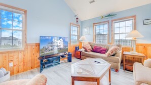 A spacious living room with a large TV, wooden walls, and a view of the sea through large windows. - Oceanview Home with Private Pool, Hot Tub, and Nearby Community Beach Access (Rodanthe)