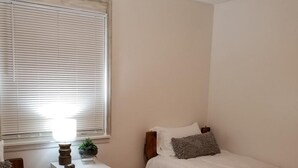 Une chambre avec deux lits simples, une table de chevet avec une lampe, une petite plante et une fenêtre avec des stores.