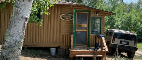 Una cabaña de madera pequeña con una puerta verde, una placa con el nombre 'Moose' y un vehículo estacionado cerca.