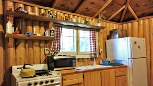 Una cocina con armarios de madera, una estufa, un microondas, un refrigerador y estantes con diversos objetos.