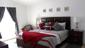 Ein ordentlich eingerichtetes Schlafzimmer mit einem großen Bett, Nachttischen, einem Sessel und einem Fenster mit Vorhängen.