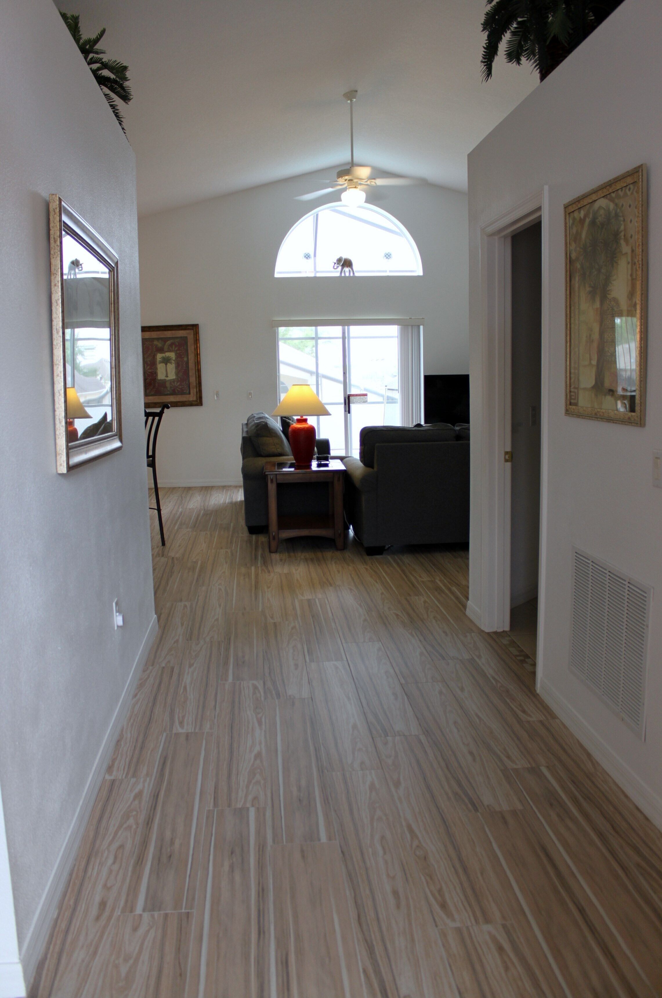 A hallway with a wooden floor, a ceiling fan, a window with a view, and a door leading to another room.