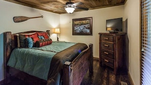 A bedroom with a wooden ceiling, a bed with a green bedspread and multiple pillows, a wooden dresser, a television, and a framed picture on the wall.