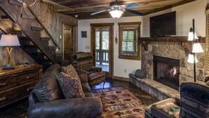 A rustic living room with a stone fireplace, leather sofa, patterned rug, and a television mounted above the fireplace.