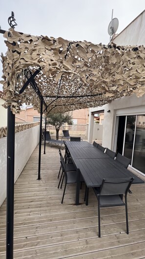 An outdoor patio with a wooden deck, a long table with chairs, and a decorative canopy.