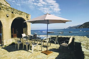 Een stenen gebouw met een tafel en stoelen onder een parasol, met uitzicht op de zee en boten.