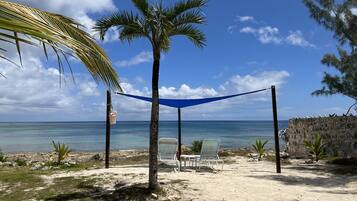 A beach with a hammock, two chairs, a small table, and a thatched structure.