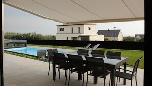 Un patio exterior con piscina, un comedor techado con sillas y vista a una casa moderna al fondo.