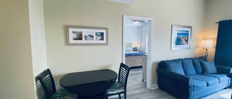 Una sala de estar con un sofá azul, una mesa de comedor con dos sillas, y un pasillo que conduce a un dormitorio con una cama y una mesita de noche.