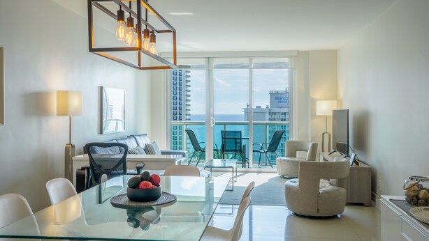 Un salon moderne avec une table à manger en verre, un canapé et un téléviseur. Il y a un balcon avec vue sur la ville et l’océan.
