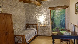 Une chambre rustique avec un lit en bois, des murs en pierre, une table en bois avec une chaise, et une fresque représentant une forêt sur le mur.
