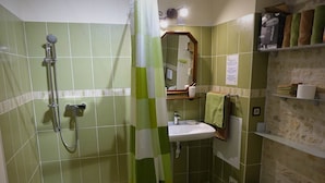 Une salle de bain avec des carreaux verts, une douche équipée d’un pommeau de douche amovible, un lavabo, un miroir et un petit tabouret.