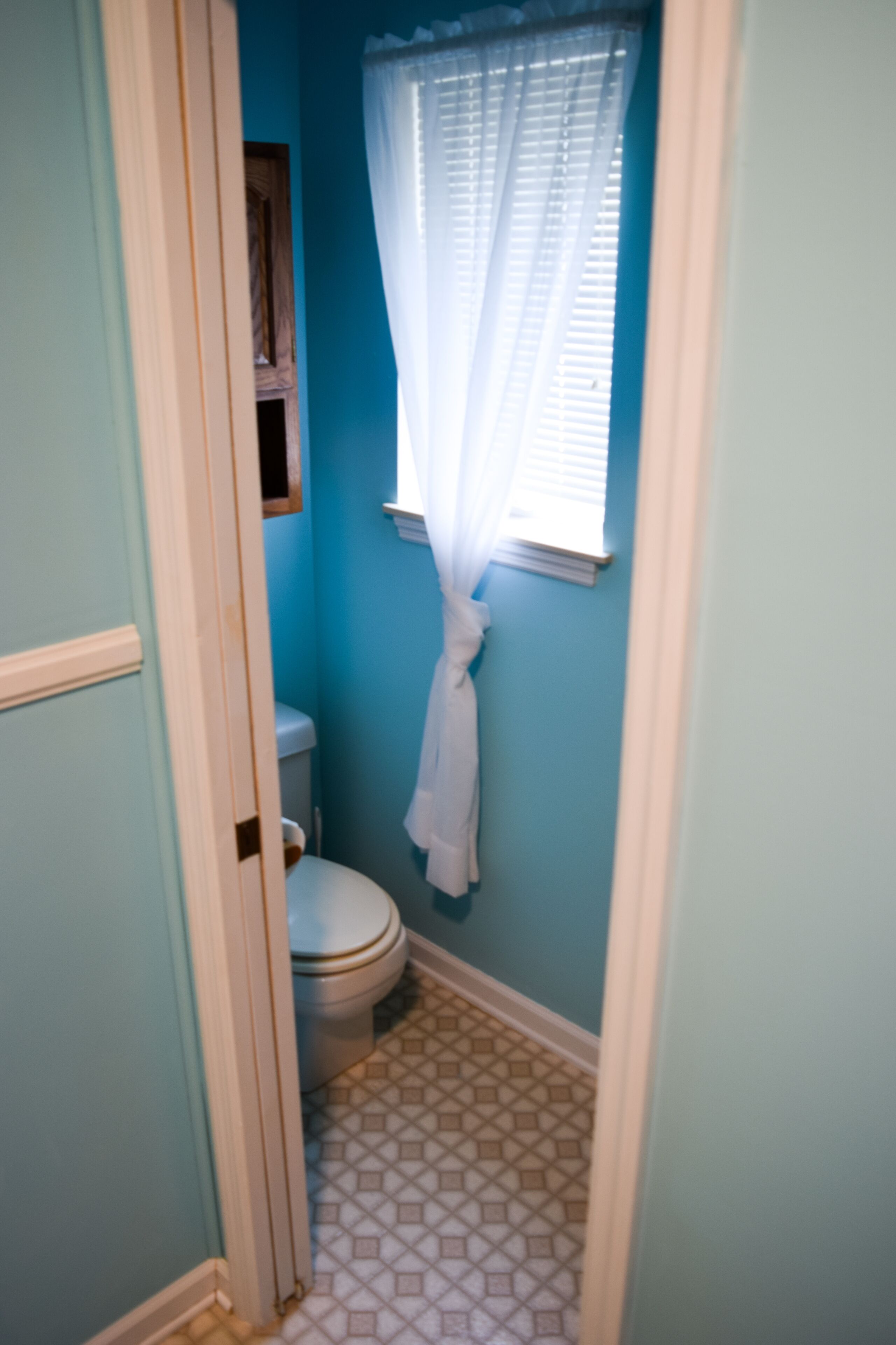 A small bathroom with a toilet, a window with a white curtain, and a tiled floor.