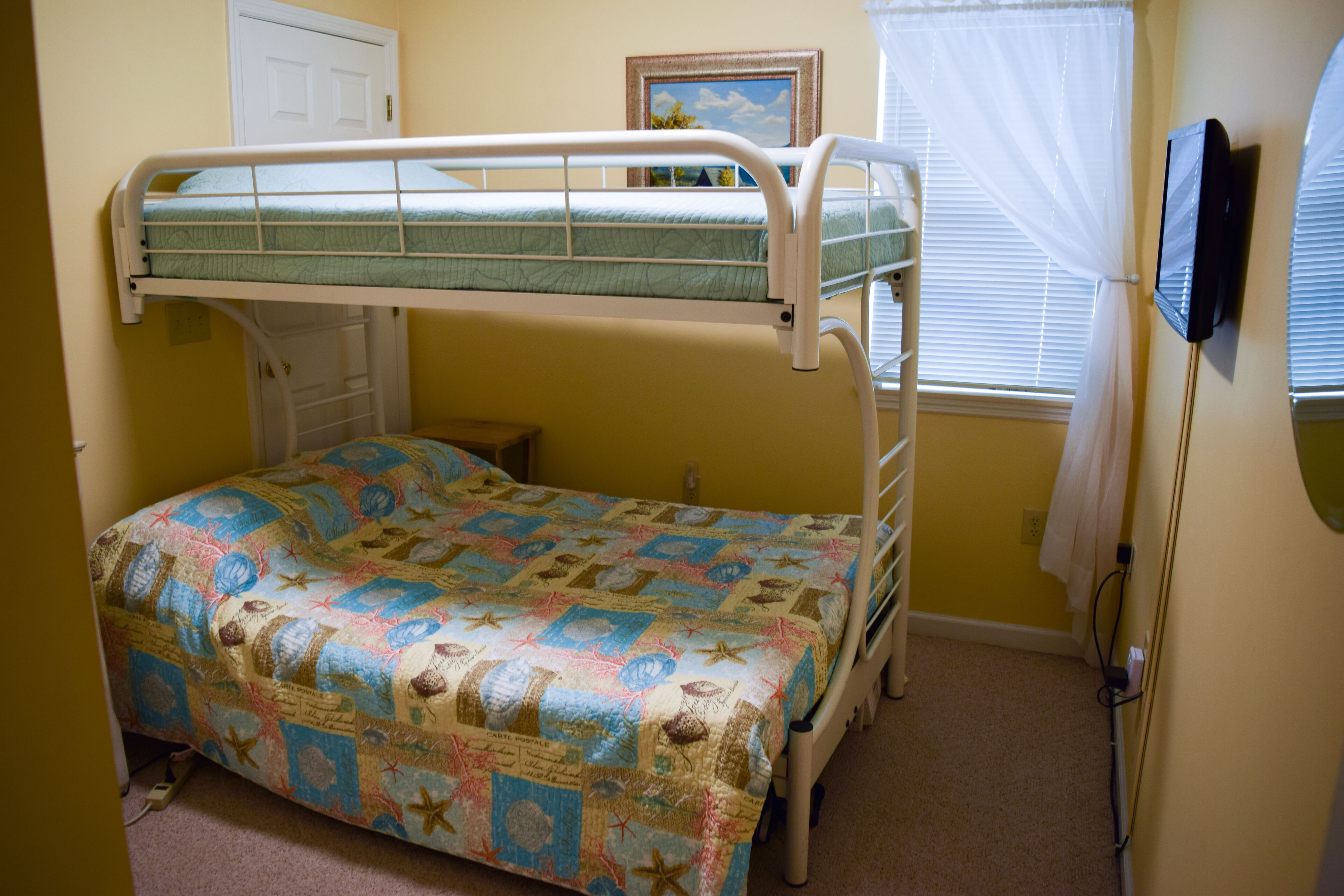 A bunk bed with a quilted bedspread, a bedside table, a window with curtains, and a wall-mounted TV.