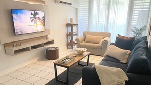 A modern living room with a flat-screen TV, a sofa, a coffee table, and a bookshelf. - Luquillo Beach Getaway – Cozy Coastal Condo Steps from  Playa Azul (Luquillo)