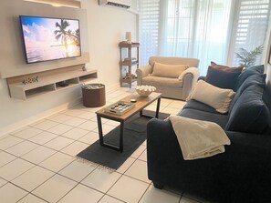 A modern living room with a flat-screen TV, a sofa, a coffee table, and a bookshelf. - Luquillo Beach Getaway – Cozy Coastal Condo Steps from  Playa Azul (Luquillo)