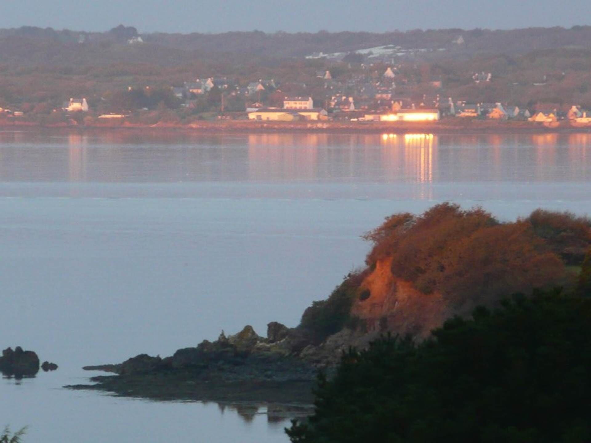Une vue côtière sur une étendue d’eau calme, des affleurements rocheux et une ville lointaine dont les bâtiments et les lumières se reflètent sur l’eau.