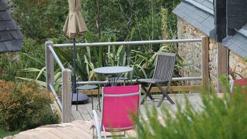 Une terrasse en bois avec une table et des chaises, entourée de verdure et d’un bâtiment en pierre.