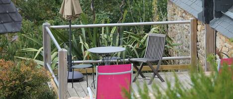 Eine Holzterrasse mit Tisch und Stühlen, umgeben von Grünpflanzen und einem Steinhaus.