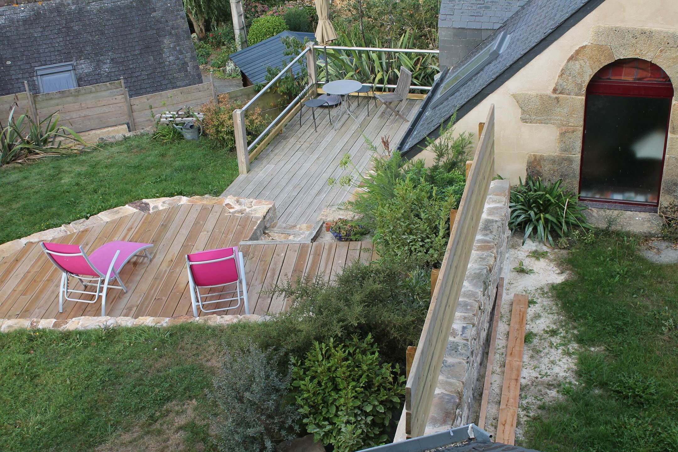 Eine Holzterrasse mit zwei rosafarbenen Stühlen und einem Tisch, umgeben von Grünpflanzen und einer Steinmauer.