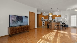 A spacious living area with a large flat-screen TV, wooden flooring, and a kitchen with wooden cabinets and a central island. - Yellowstone Mountain View  (Gardiner)